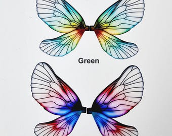 Op de afbeelding: Twee paar kleurrijke vlindervleugels. Eén paar is groen, geel en blauw, het andere is rood, roze en blauw. De vleugels zijn gelabeld met "Green" en "Blue" in zwarte tekst. De vleugels hebben een zwart midden en zijn gemaakt van een doorschijnend materiaal.