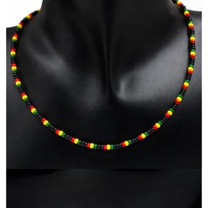 May include: A beaded necklace with a Rastafarian color scheme. The necklace features small, round beads in black, red, yellow, and green, strung together to form a necklace. The necklace is displayed on a black mannequin.