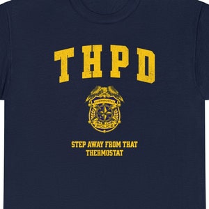 以下が含まれることがあります： 紺色のTシャツ。黄色で「THPD」の文字、黄色の警察バッジ、そして「STEP AWAY FROM THAT THERMOSTAT」というフレーズがプリントされています。クルーネックのクラシックなデザインです。