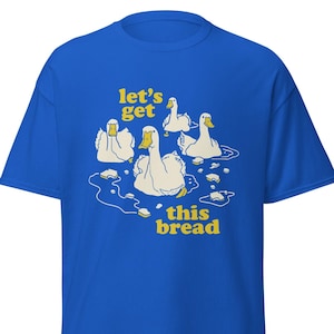 Può includere: T-shirt blu reale con grafica di anatre dei cartoni animati nell'acqua, con la scritta gialla "let's get this bread". Le anatre sono bianche con becco e zampe gialle. La maglietta è uno stile casual unisex.