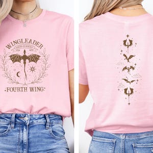 May include: Pink t-shirt with a brown graphic of a dragon and the text "WINGLEADER RIDERS GUILD" and "FOURTH WING". The back of the shirt has a vertical line of brown dragon graphics.