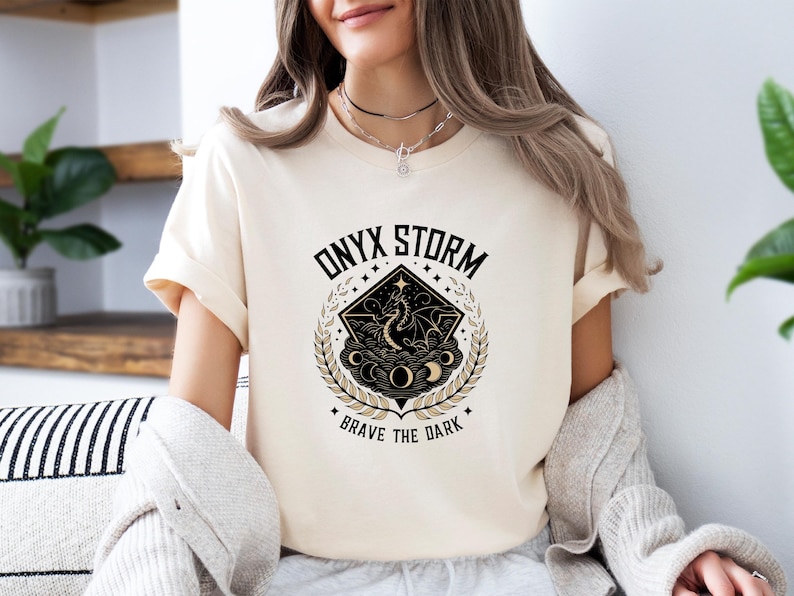 Puede incluir: Una camiseta blanca con un dise&ntilde;o gr&aacute;fico en negro y dorado que presenta un drag&oacute;n, estrellas y lunas. El texto "ONYX STORM" y "BRAVE THE DARK" est&aacute; impreso en la camiseta.
