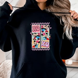 May include: Black hoodie featuring a colorful graphic of Mickey and Minnie Mouse in various romantic poses. The design includes the letters "XO" and a pink and white checkered border. The hoodie has a drawstring hood and a front pocket.