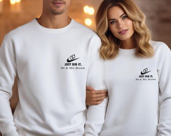 May include: Two white crewneck sweatshirts featuring a black graphic on the left chest. The graphic includes a swoosh symbol with two interlocking rings above it, followed by the text "JUST DID IT." and "Mr & Mrs Brown" in black lettering.