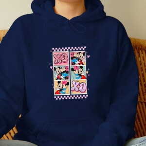 May include: Navy blue hoodie featuring a graphic of Mickey and Minnie Mouse in a comic strip style. The design includes the letters "XO" and pink and black checkered borders. The hoodie is made of a soft material.