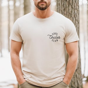 Könnte beinhalten: Ein beiges T-Shirt mit einem schwarzen Aufdruck, der in kursivschrift "Cool Uncles Club" lautet.