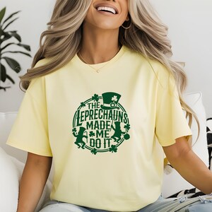 May include: A light yellow t-shirt with a green graphic that reads "THE LEPRECHAUNS MADE ME DO IT." The design includes a leprechaun hat, shamrocks, and leprechaun figures. The shirt is a casual, oversized fit.