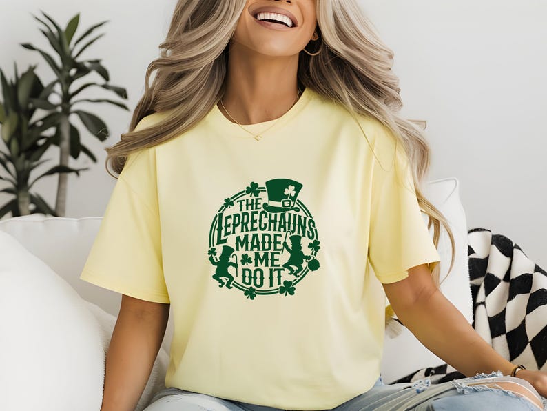 May include: A light yellow t-shirt with a green graphic that reads "THE LEPRECHAUNS MADE ME DO IT." The design includes a leprechaun hat, shamrocks, and leprechaun figures. The shirt is a casual, oversized fit.