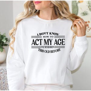 May include: A white long-sleeve crewneck sweatshirt featuring black text on the front that reads I DON'T KNOW HOW TO ACT MY AGE I'VE NEVER BEEN THIS OLD BEFORE, with horizontal lines framing the text above and below the middle phrase.