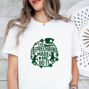 May include: White t-shirt with a green graphic. The graphic features a leprechaun hat, shamrocks, and the text "THE LEPRECHAUNS MADE ME DO IT" in a circular design. The shirt is a casual style.