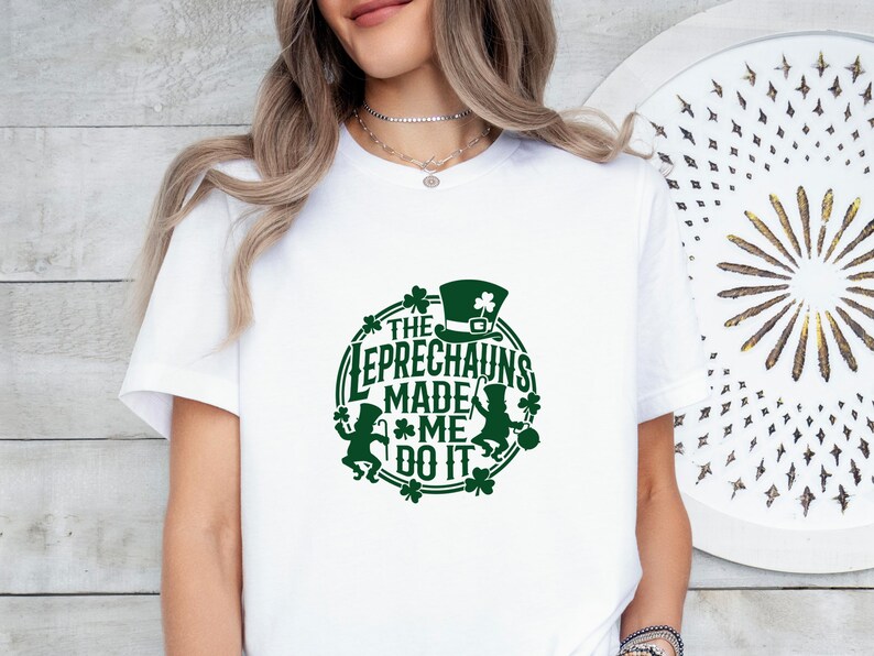 May include: White t-shirt with a green graphic. The graphic features a leprechaun hat, shamrocks, and the text "THE LEPRECHAUNS MADE ME DO IT" in a circular design. The shirt is a casual style.