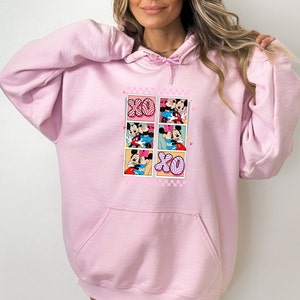 May include: Light pink hoodie with a front graphic featuring Mickey and Minnie Mouse in various romantic poses. The graphic includes the letters "XO" in a checkered pattern. The hoodie has a drawstring hood and a front pocket.
