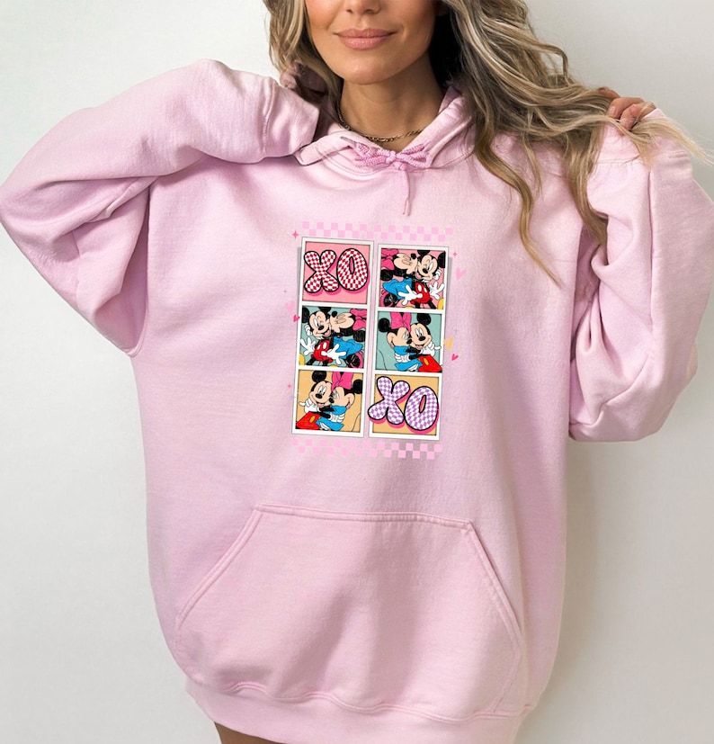 May include: Light pink hoodie with a front graphic featuring Mickey and Minnie Mouse in various romantic poses. The graphic includes the letters "XO" in a checkered pattern. The hoodie has a drawstring hood and a front pocket.