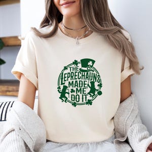 May include: Cream-colored t-shirt with a green graphic. The graphic features the text "THE LEPRECHAUNS MADE ME DO IT" in a circular design, along with leprechaun figures, a hat, and shamrocks. The shirt is a casual style.