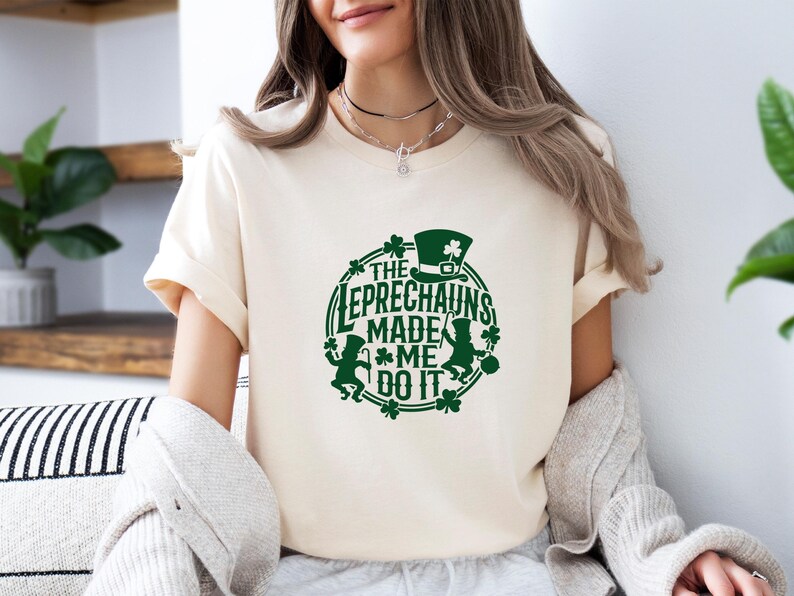 May include: Cream-colored t-shirt with a green graphic. The graphic features the text "THE LEPRECHAUNS MADE ME DO IT" in a circular design, along with leprechaun figures, a hat, and shamrocks. The shirt is a casual style.