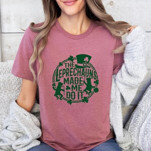 May include: A dusty rose t-shirt with a green graphic design. The design features a leprechaun hat, shamrocks, and the text "The Leprechauns Made Me Do It" in a circular arrangement. The shirt is made of soft material.