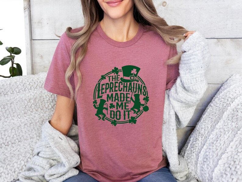 May include: A dusty rose t-shirt with a green graphic design. The design features a leprechaun hat, shamrocks, and the text "The Leprechauns Made Me Do It" in a circular arrangement. The shirt is made of soft material.