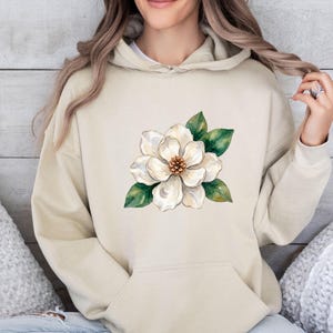 Op de afbeelding: Een beige hoodie met een grote, centraal geplaatste aquarelillustratie van een witte magnolia-bloem met groene bladeren en een bruin hart. Het kledingstuk is voorzien van een kangoeroezak, lange mouwen en een capuchon met trekkoord.