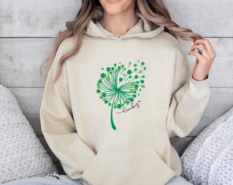 May include: A beige hooded sweatshirt featuring a green graphic of a dandelion where the seeds are replaced by small four-leaf clovers. The word lucky is written in black cursive script below the graphic, accompanied by a small black heart.