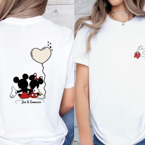 May include: White t-shirt with a cartoon illustration of Mickey and Minnie Mouse. The back of the shirt shows the couple with a heart-shaped balloon. The front has a small design of two rabbits forming a heart.