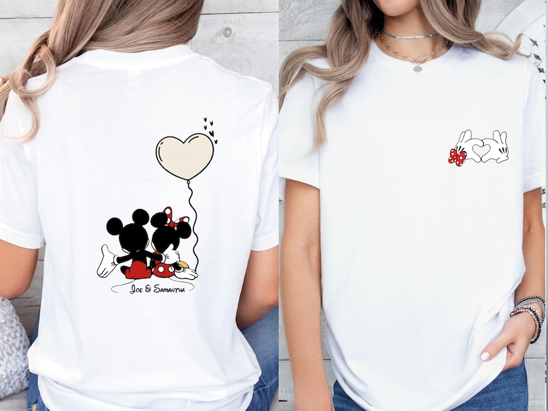 May include: White t-shirt with a cartoon illustration of Mickey and Minnie Mouse. The back of the shirt shows the couple with a heart-shaped balloon. The front has a small design of two rabbits forming a heart.