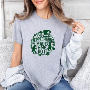 May include: A light gray t-shirt with a green graphic that says "The Leprechauns Made Me Do It." The design includes a leprechaun hat, shamrocks, and leprechaun figures. The shirt is short-sleeved.