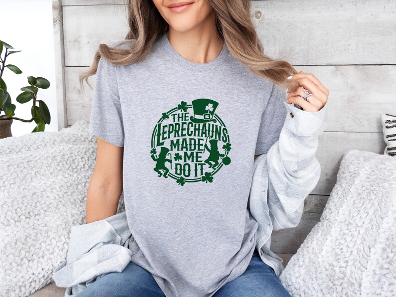 May include: A light gray t-shirt with a green graphic that says "The Leprechauns Made Me Do It." The design includes a leprechaun hat, shamrocks, and leprechaun figures. The shirt is short-sleeved.