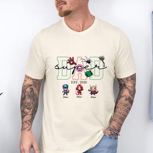 May include: Cream-coloured t-shirt with the word "DAD" in green and red block letters, with the word "SUPER" in a script font. The shirt features superhero-themed graphics and the text "EST. 2026".