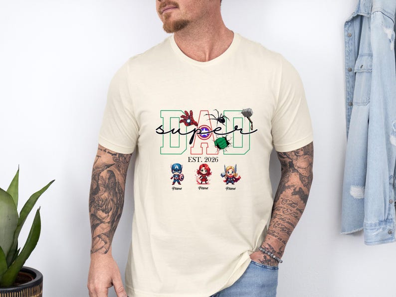 May include: Cream-coloured t-shirt with the word "DAD" in green and red block letters, with the word "SUPER" in a script font. The shirt features superhero-themed graphics and the text "EST. 2026".