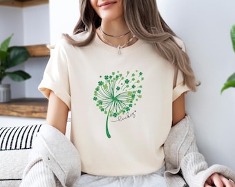 May include: A cream-colored short-sleeve t-shirt featuring a green graphic of a dandelion where the seeds are shaped like four-leaf clovers. The word lucky is written in black cursive script next to the dandelion design on the front of the shirt.