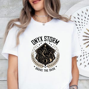 Puede incluir: Camiseta blanca con un dise&ntilde;o gr&aacute;fico en negro y dorado que representa un drag&oacute;n, nubes y estrellas. El texto "ONYX STORM" y "BRAVE THE DARK" est&aacute; impreso en la camiseta.