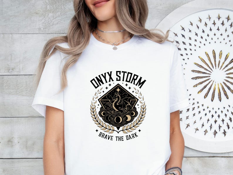 Puede incluir: Camiseta blanca con un dise&ntilde;o gr&aacute;fico en negro y dorado que representa un drag&oacute;n, nubes y estrellas. El texto "ONYX STORM" y "BRAVE THE DARK" est&aacute; impreso en la camiseta.
