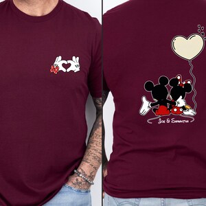 May include: Burgundy t-shirt with a white hand-heart design and a red bow. The back of the shirt shows a cartoon couple with a heart-shaped balloon and the text "Joe & Samantha".