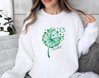 May include: A white long-sleeve crewneck sweatshirt featuring a graphic of a dandelion where the seeds are replaced by small green four-leaf clovers. The word lucky is written in black cursive script below the green clover dandelion design.