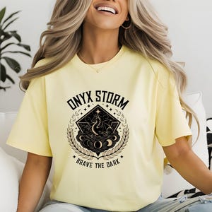 Puede incluir: Una camiseta amarilla con un dise&ntilde;o gr&aacute;fico negro que presenta un drag&oacute;n, estrellas y el texto "ONYX STORM BRAVE THE DARK".