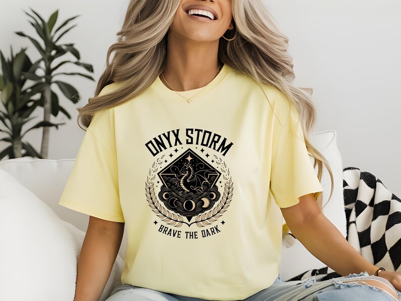 Puede incluir: Una camiseta amarilla con un dise&ntilde;o gr&aacute;fico negro que presenta un drag&oacute;n, estrellas y el texto "ONYX STORM BRAVE THE DARK".