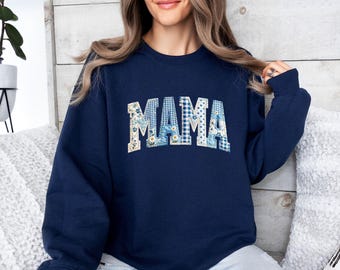 May include: A navy blue crewneck sweatshirt featuring the word MAMA in large, arched block letters. The letters are filled with a patchwork design of blue and white floral patterns, gingham checks, and small polka dots in various shades of blue.