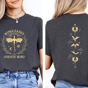 May include: A dark gray t-shirt with a gold graphic of a dragon with the text "WINGLEADER RIDERS GUARDIAN FOURTH WING" on the front. The back of the shirt has a vertical row of gold dragon graphics.