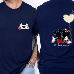 May include: Navy blue t-shirt with a Mickey and Minnie Mouse design. The front shows a heart made of white gloved hands with a red bow. The back features the couple with a heart balloon and the names "Joe & Samantha".