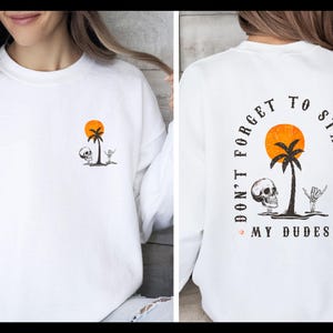May include: A white crewneck sweatshirt featuring a small graphic on the front left chest and a larger version on the back. The design includes an orange sun, a black palm tree, a skull, and a skeleton hand with the text DON'T FORGET TO STAY SALTY MY DUDES.