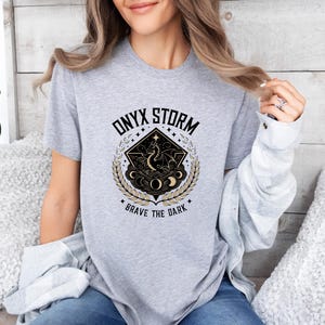 Puede incluir: Camiseta gris con un dise&ntilde;o gr&aacute;fico en negro y dorado que representa un drag&oacute;n, estrellas y lunas. El texto "ONYX STORM" y "BRAVE THE DARK" est&aacute; impreso en la camiseta.