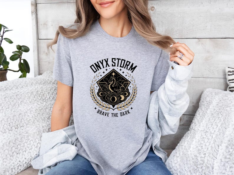 Puede incluir: Camiseta gris con un dise&ntilde;o gr&aacute;fico en negro y dorado que representa un drag&oacute;n, estrellas y lunas. El texto "ONYX STORM" y "BRAVE THE DARK" est&aacute; impreso en la camiseta.