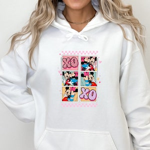 May include: White hoodie featuring a colorful graphic of Mickey and Minnie Mouse in various romantic poses. The design includes the letters "XO" and a pink checkered border, perfect for Valentine's Day or Disney fans.