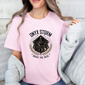 Puede incluir: Camiseta rosa con un dise&ntilde;o gr&aacute;fico en negro y dorado que representa un drag&oacute;n, lunas y estrellas. El texto "ONYX STORM" y "BRAVE THE DARK" est&aacute; impreso en la camiseta.
