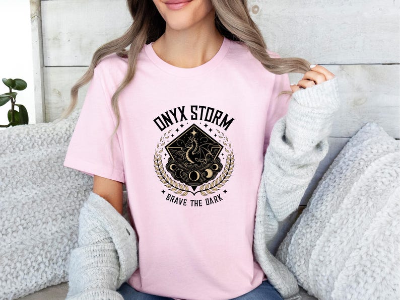 Puede incluir: Camiseta rosa con un dise&ntilde;o gr&aacute;fico en negro y dorado que representa un drag&oacute;n, lunas y estrellas. El texto "ONYX STORM" y "BRAVE THE DARK" est&aacute; impreso en la camiseta.