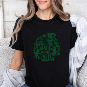 May include: Black t-shirt with a green graphic design. The design features a leprechaun hat, shamrocks, and the text "THE LEPRECHAUNS MADE ME DO IT" in a circular arrangement. A festive St. Patrick's Day themed tee.