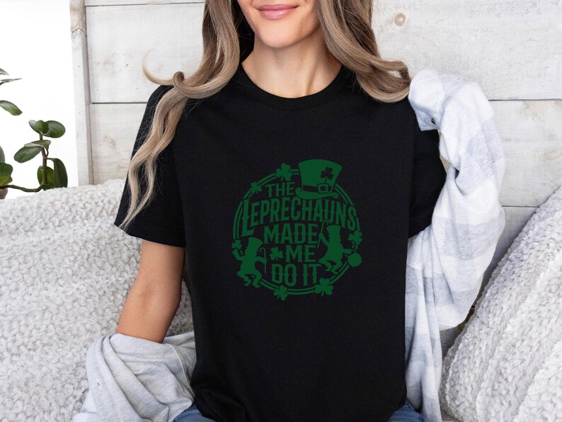 May include: Black t-shirt with a green graphic design. The design features a leprechaun hat, shamrocks, and the text "THE LEPRECHAUNS MADE ME DO IT" in a circular arrangement. A festive St. Patrick's Day themed tee.