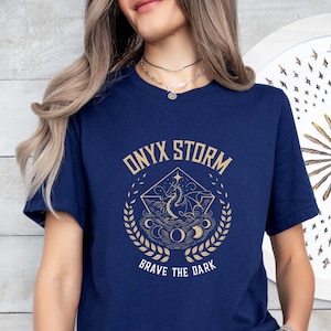Puede incluir: Una camiseta azul marino con un gr&aacute;fico dorado de un drag&oacute;n y el texto "ONYX STORM BRAVE THE DARK".