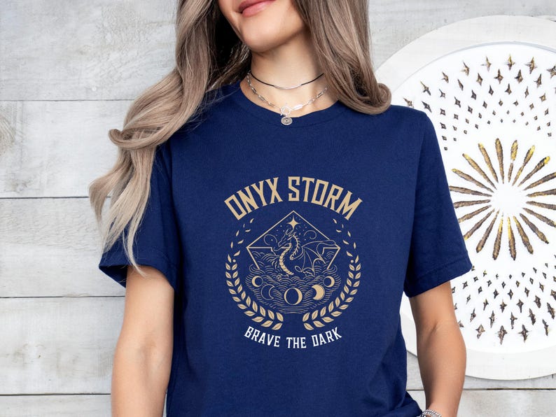 Puede incluir: Una camiseta azul marino con un gr&aacute;fico dorado de un drag&oacute;n y el texto "ONYX STORM BRAVE THE DARK".