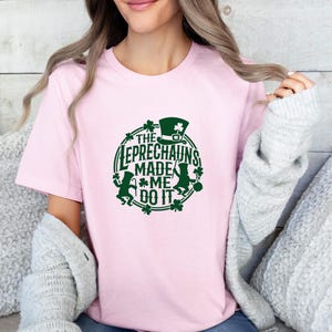 May include: A light pink t-shirt with a green graphic that says "THE LEPRECHAUNS MADE ME DO IT." The design includes a leprechaun, a hat, and shamrocks. The shirt is a crew neck style.